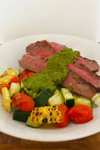 Chimichurri sauce on top of grilled steak, zucchini, cherry tomatoes, corn, and rice in a bowl.