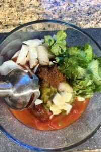 Immersion blender blending all of the homemade salsa ingredients in a clear glass bowl.